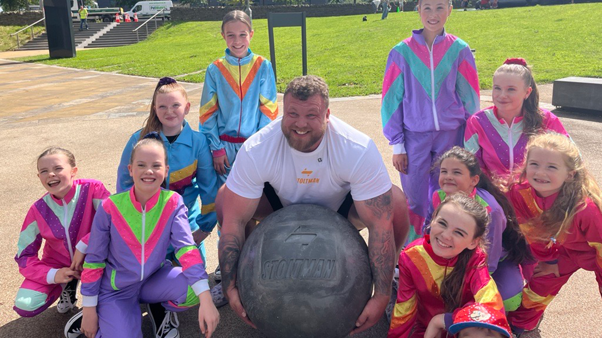 Tom Stoltman carrying a big stone surrounded by young dancers in track suits